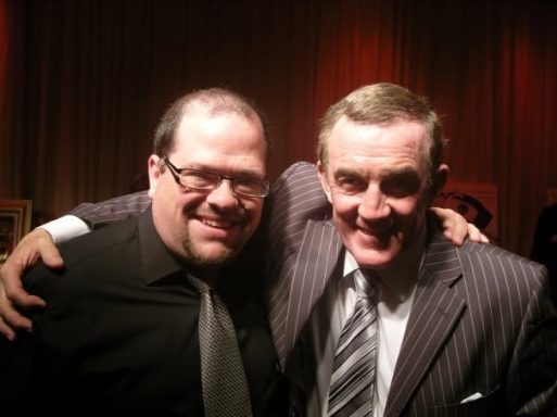 Two smiling men pose closely together, one in a black shirt and the other in a pinstripe suit.