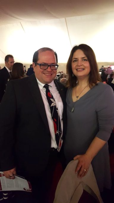 Two people, a man in a suit and a woman in a grey dress, smiling at an event.