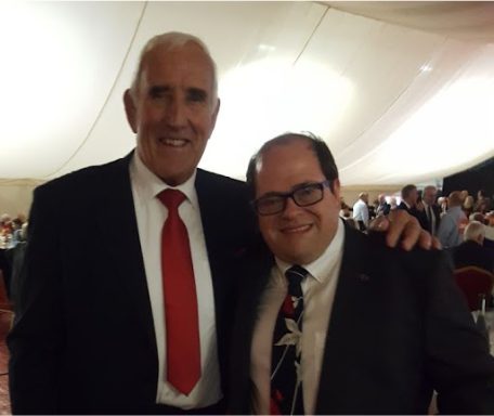 Two men, one in a black suit with a red tie and the other in formal attire, smiling together.