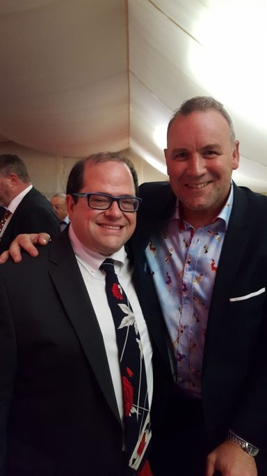 Two men smiling and posing for a photo, one in a patterned shirt and the other in glasses.