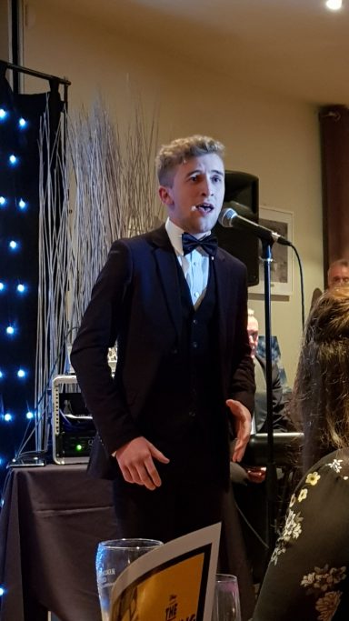 A young man in a tuxedo singing at a formal event with a microphone.