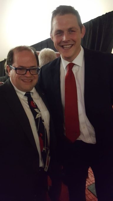 Two men posing together, one in a suit with a red tie and the other in a patterned tie.