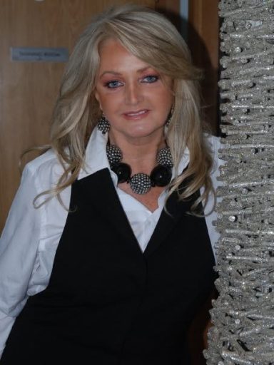 A woman with long blonde hair wearing a black vest and statement necklace, smiling.